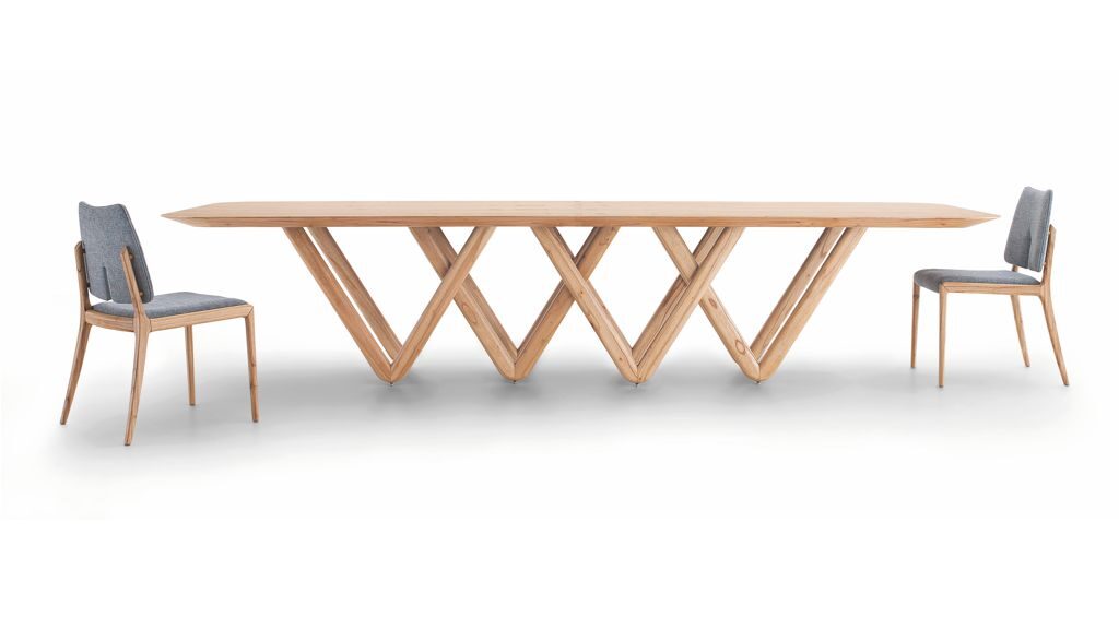 Na imagem, é possível ver um mesa com os pés parecidos com a letra W. Nas pontas externas, é possível visualizar duas cadeiras que, assim como a mesa, possuem madeira maciça em sua composição. 