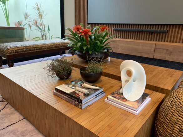 Na imagem, é possível ver um vaso de flor vermelha e alguns objetos de decoração compondo a mesa de madeira. Ao fundo, móveis de fibra natural e ripas de madeira